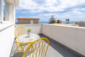 A balcony with a round table and yellow chairs, overlooking a coastal area with buildings and palm trees.