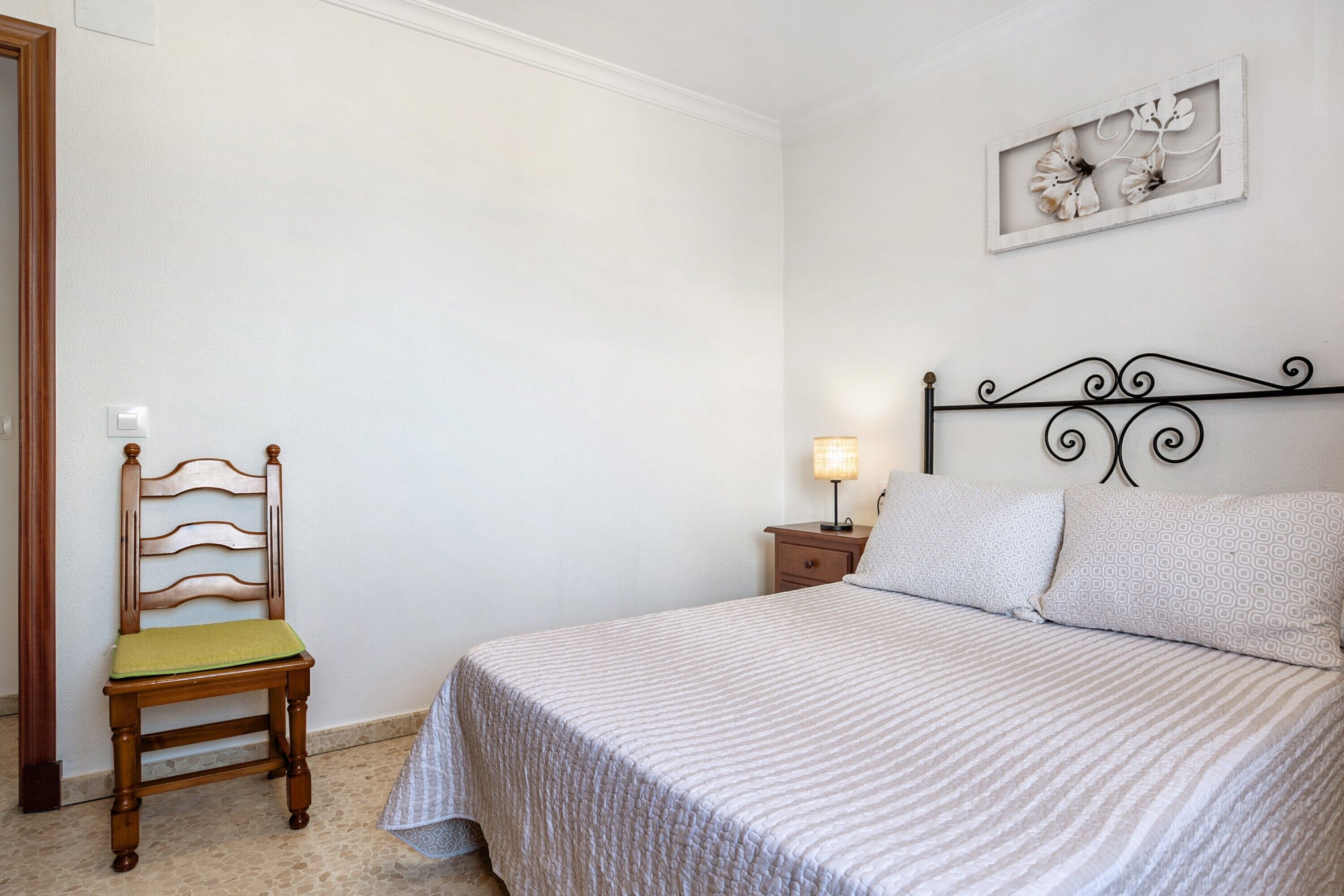Une chambre à coucher comprenant un lit, une chaise, une table de chevet, une lampe et une œuvre d’art accrochée au mur.
