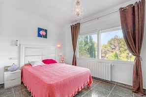 Une chambre à coucher comprenant un lit, une table de chevet, une lampe fixée au mur, une fenêtre avec des rideaux et une vue sur les arbres.