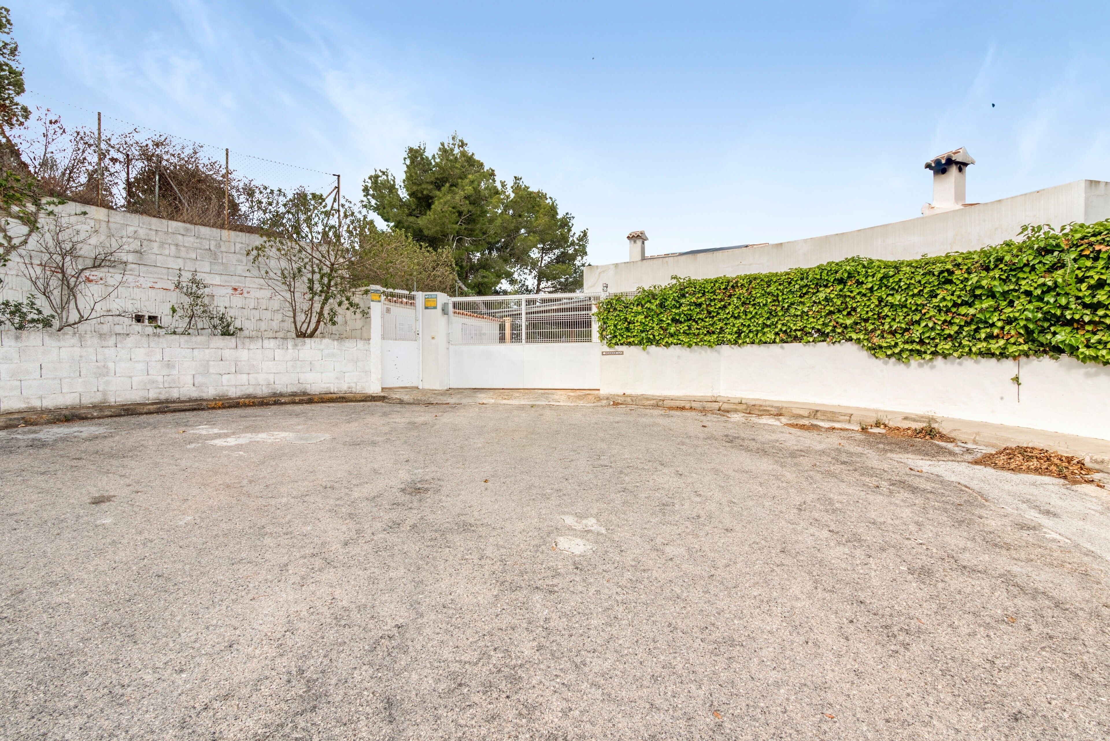 A paved courtyard with a white wall, a metal gate, and a hedge.