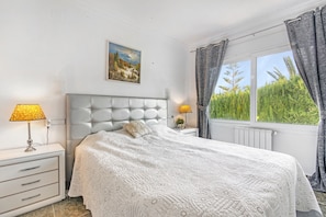 Een slaapkamer met een gestuukt hoofdeinde, een bed met een wit sprei, een nachtkastje met lampen en een raam met gordijnen.