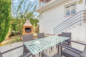 Une terrasse extérieure avec une table à plateau de verre et des chaises en osier, un barbecue en briques et un bâtiment blanc doté d’une fenêtre et d’un escalier.
