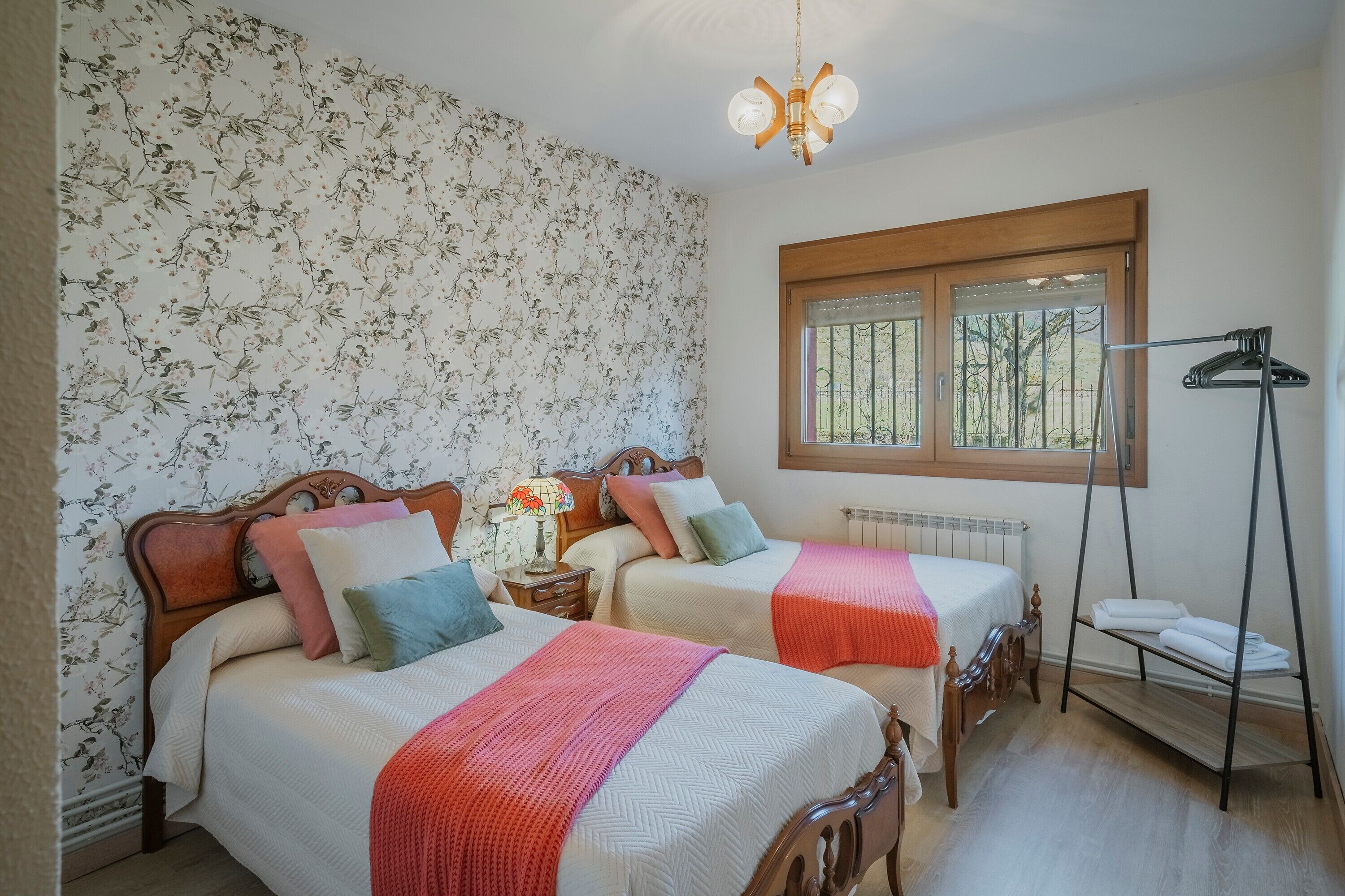 Un dormitorio con dos camas, papel tapiz floral, cabecero de madera, una lámpara de noche, un radiador, una ventana con persianas y una lámpara de pie.