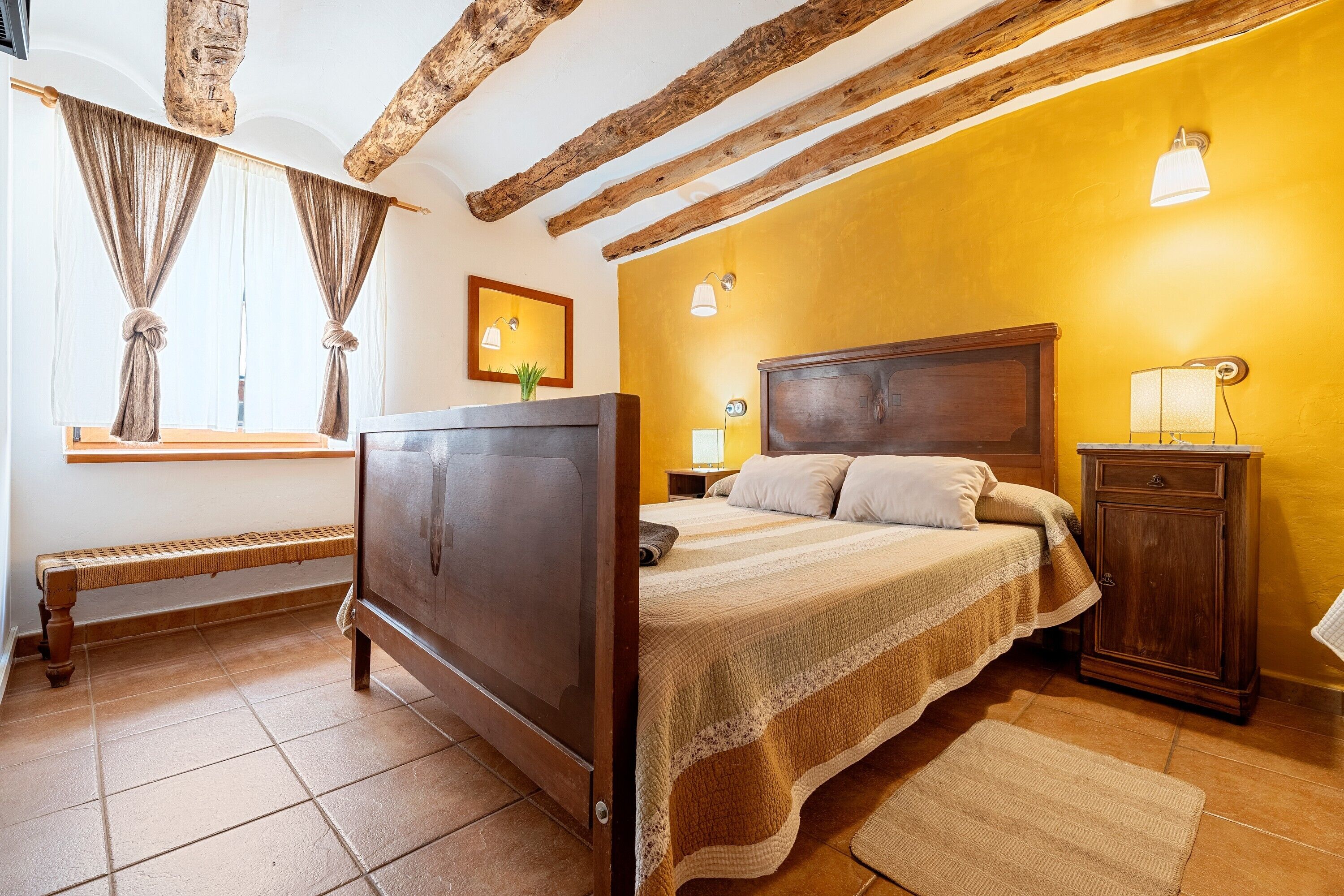 Ein Schlafzimmer mit einem hölzernen Bett, einem Nachttisch, einer Bank und einem Fenster mit Vorhängen.