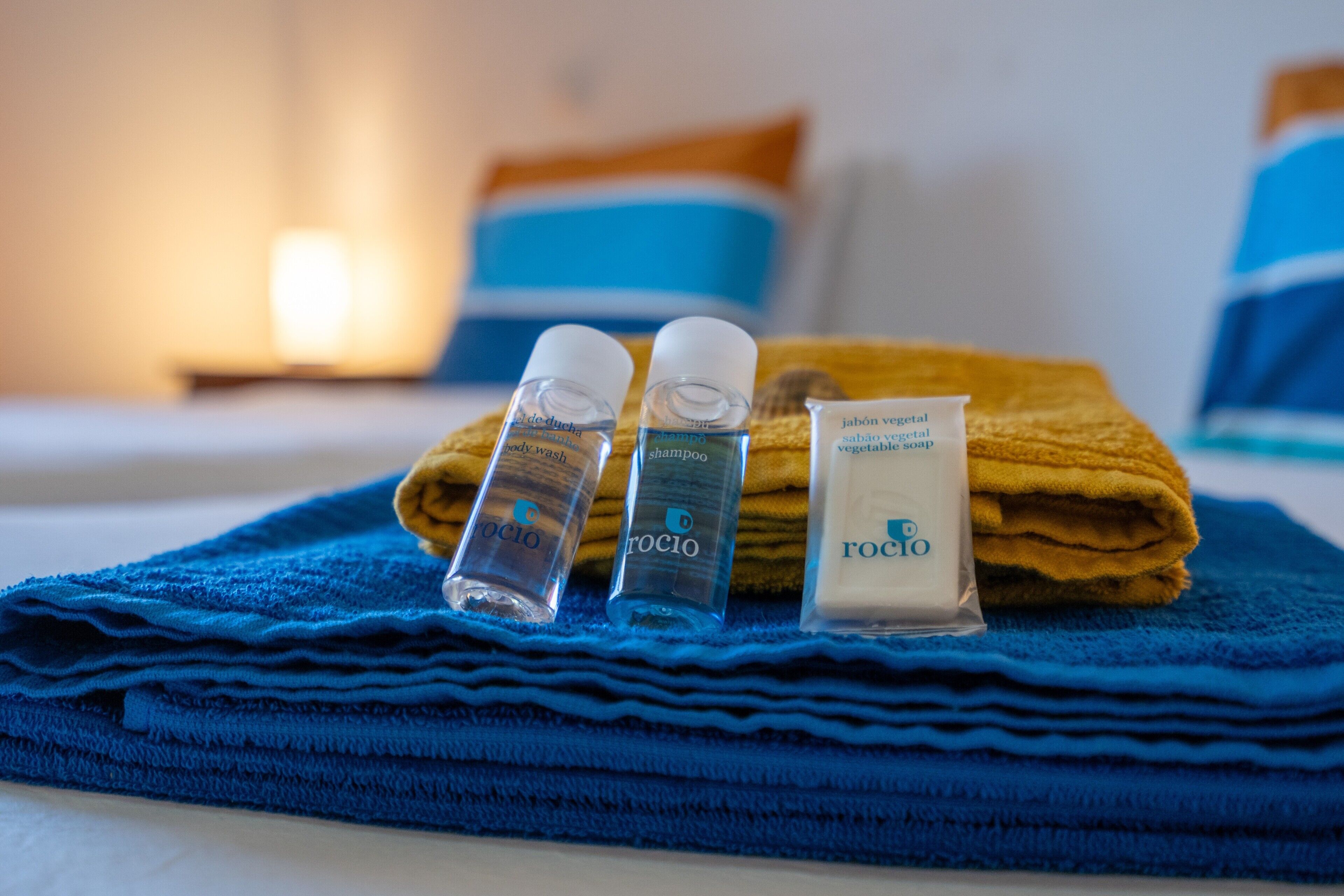 A neatly made bed with blue and orange pillows, a blue towel, and toiletries including shampoo and soap.