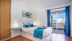 Un dormitorio con cama, mesita de noche, un cuadro colgado en la pared y un balcón con cortinas azules.