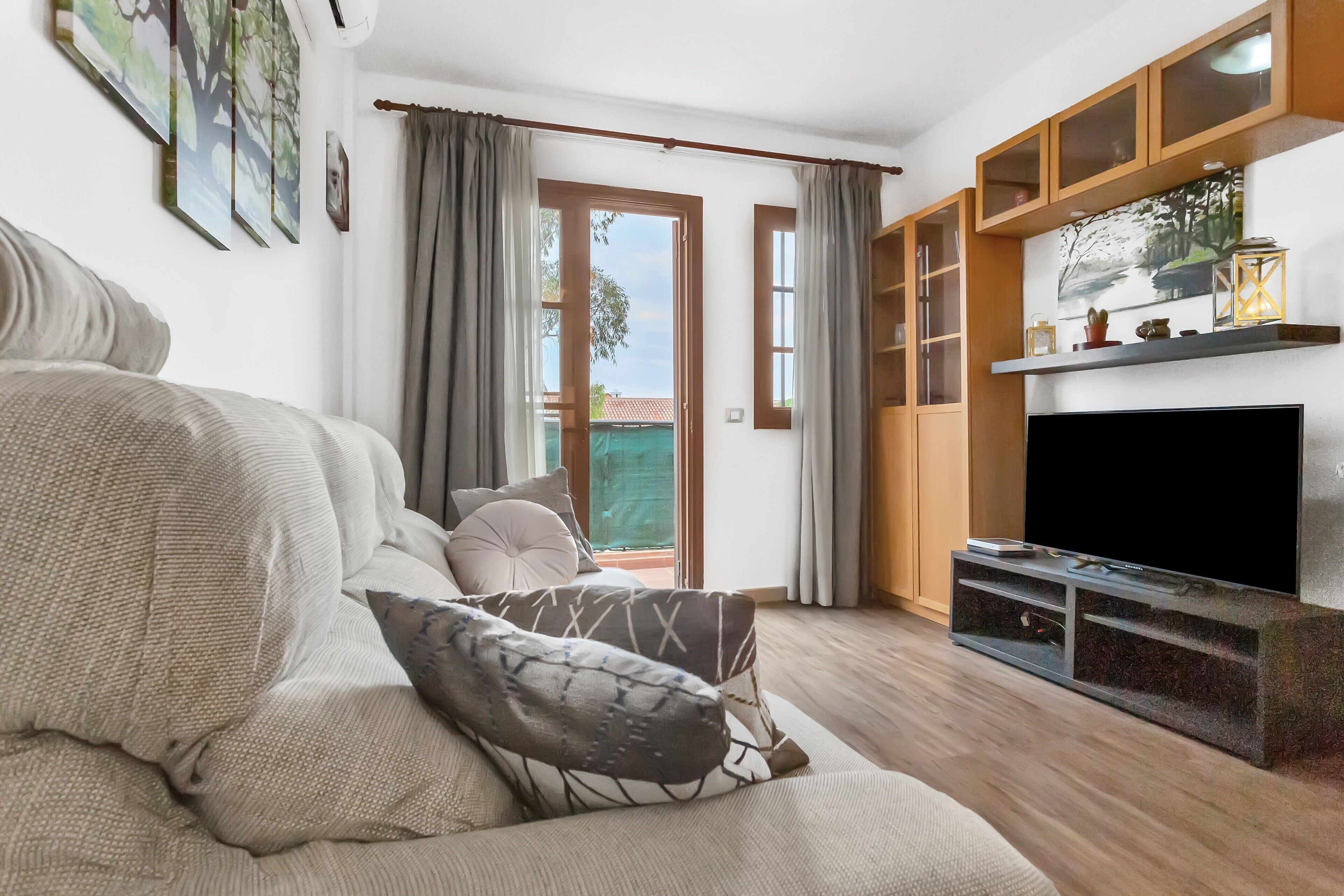 Een woonkamer met een bank, een flatscreen-tv, houten kasten en een schuifpui naar een balkon.