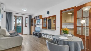 A modern living room with a sofa, a dining table with chairs, a kitchen area with cabinets, and a TV.