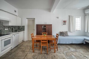 A compact kitchen with white appliances, a dining table with chairs, a wooden cabinet, and a bed with a blue bedspread.