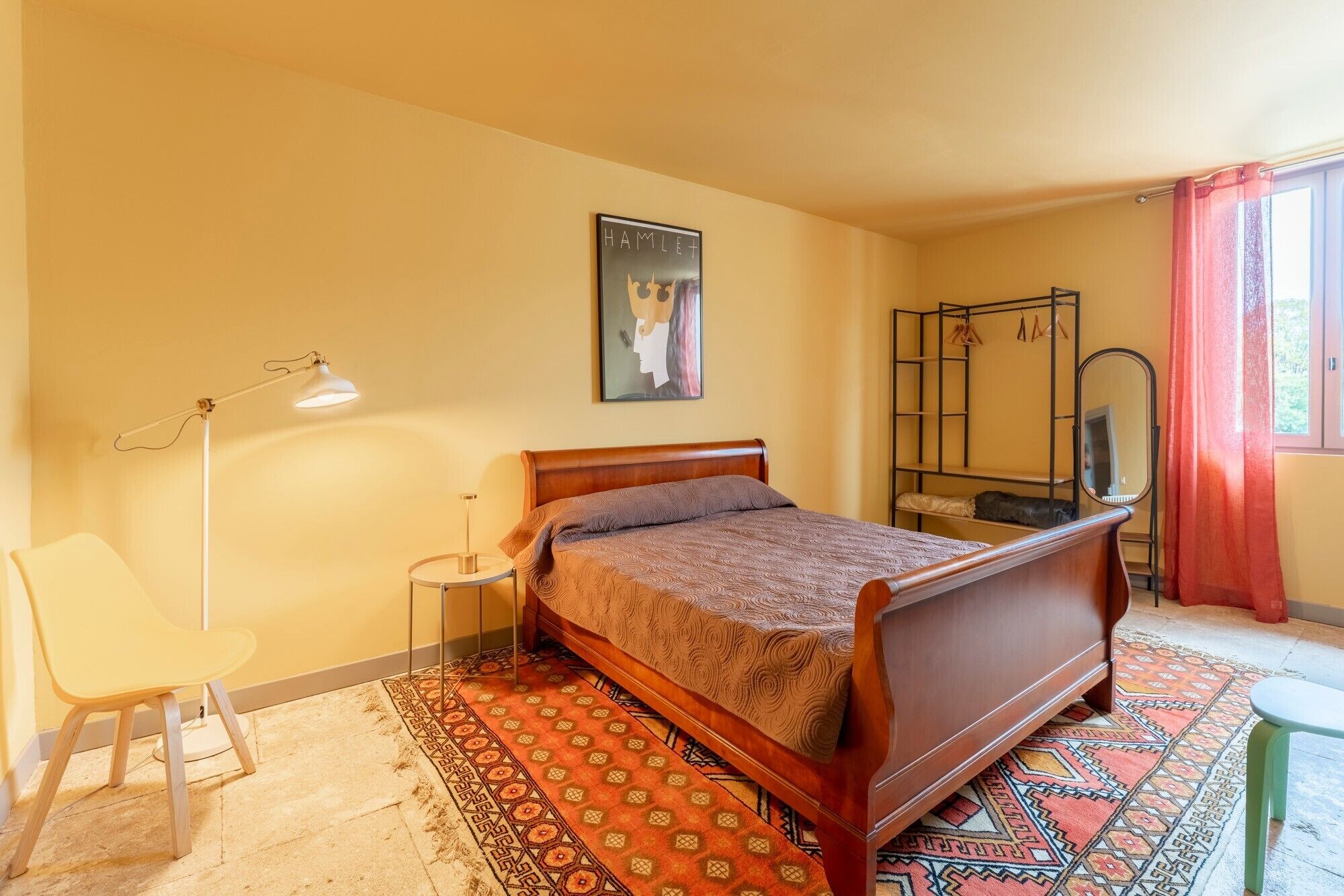 Une chambre à coucher avec un lit en bois, une lampe sur pied, un miroir fixé au mur, une affiche et un tapis à motifs.