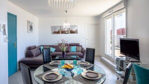 A modern living room with a sofa, a dining table set for four, a TV, and artwork on the walls. - Apartment 'Appart Villa Margaux Near the Sea' with Private Terrace and Wi-Fi (Les Sables-d'Olonne)