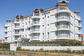 A multi-story residential building with balconies, a flat roof, and a hedge in front.