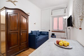 A room with a wooden wardrobe, a blue sofa, a table with fruit, and a TV on the wall.