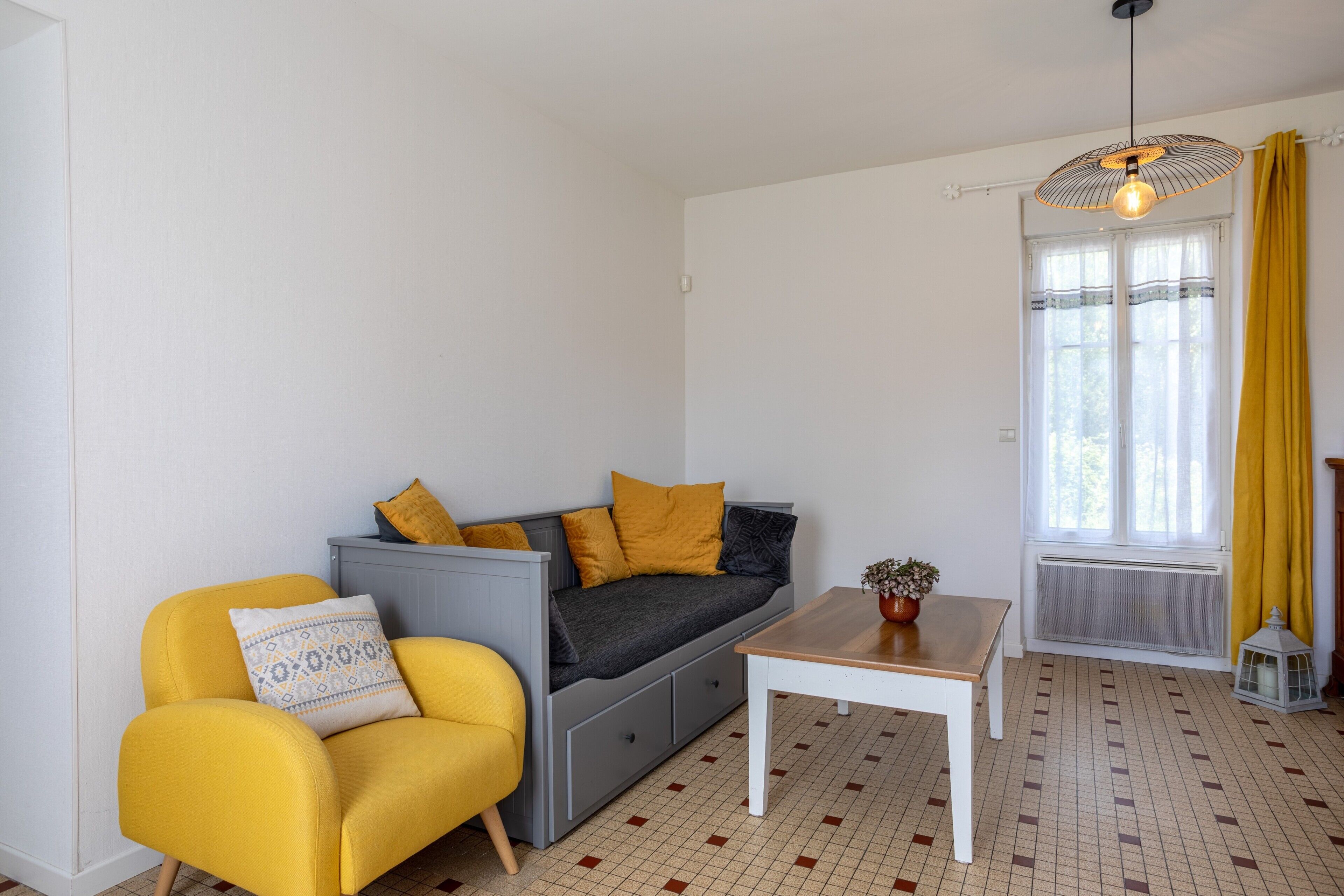 A room with a sofa, a yellow armchair, a small table, a hanging light fixture, and a window with curtains.
