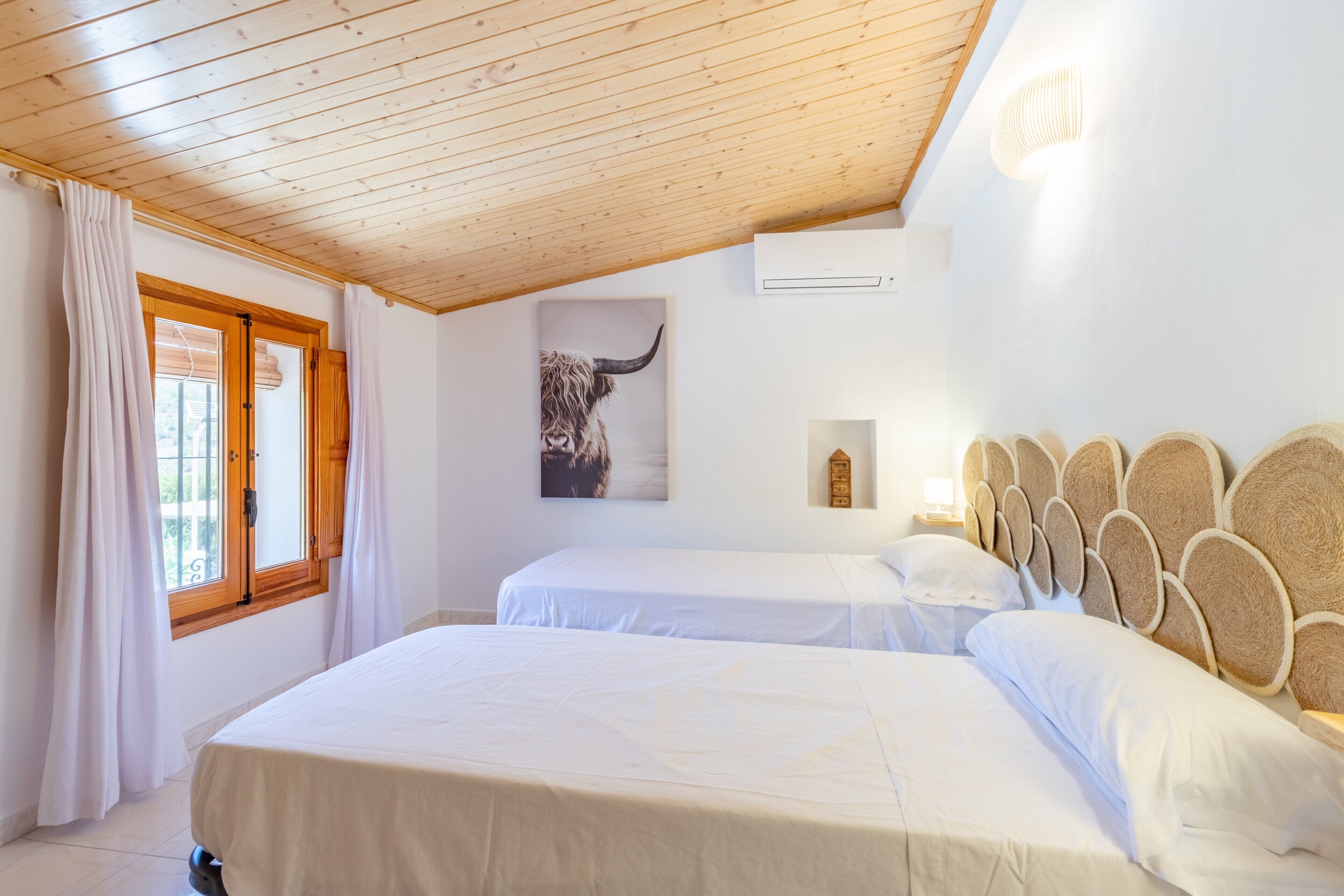 Ein Schlafzimmer mit zwei Betten, Holzboden, einem Fenster mit Vorhängen, einer Wand-Klimaanlage und einem Bild eines Stiers.