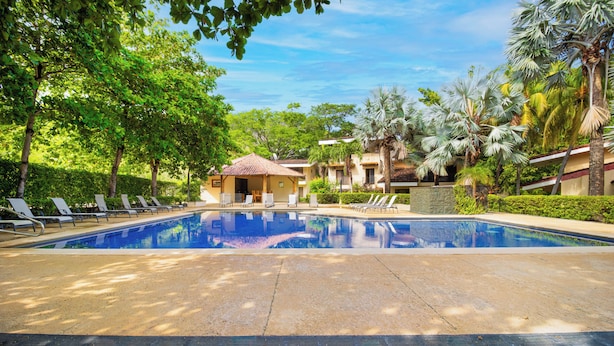 A swimming pool with lounge chairs, surrounded by palm trees and a small building with a tiled roof. - Ocotal Beach House - Family Friendly & Only a Block To The Beach! (Ocotal)