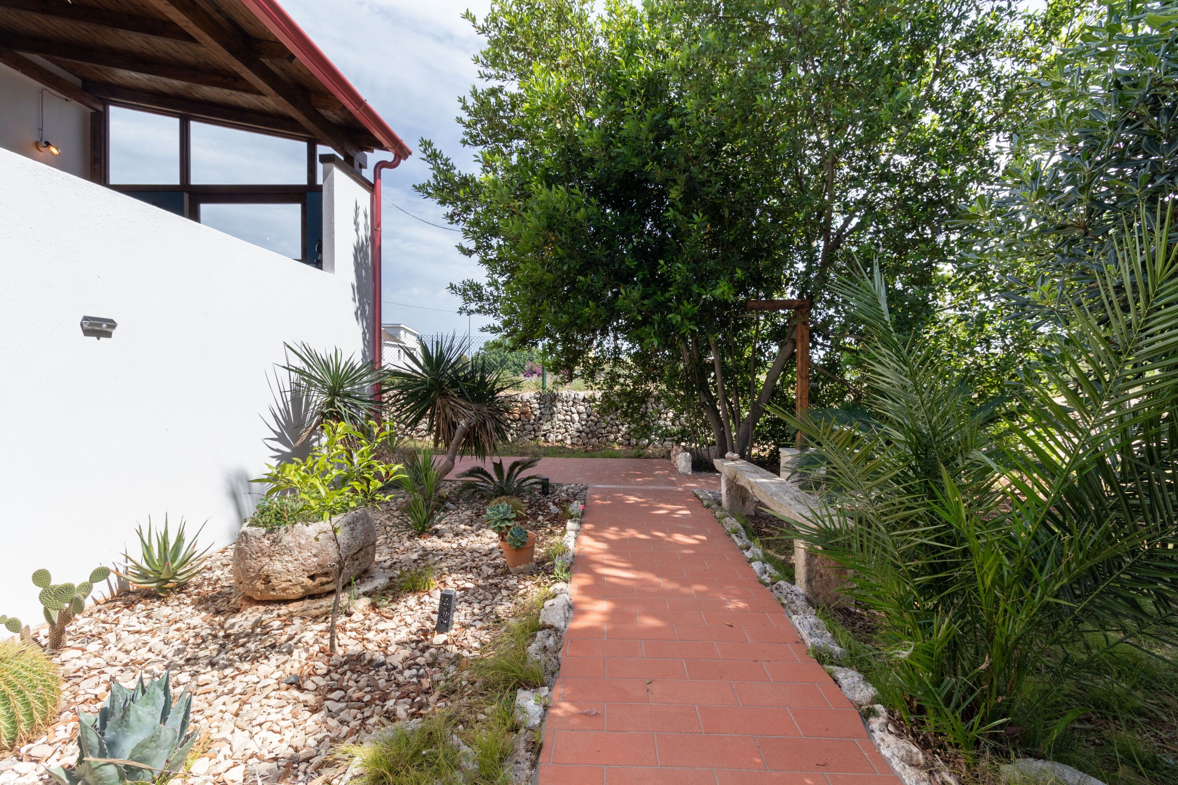 A pathway leading to a building with a white exterior, surrounded by various plants and a stone wall.