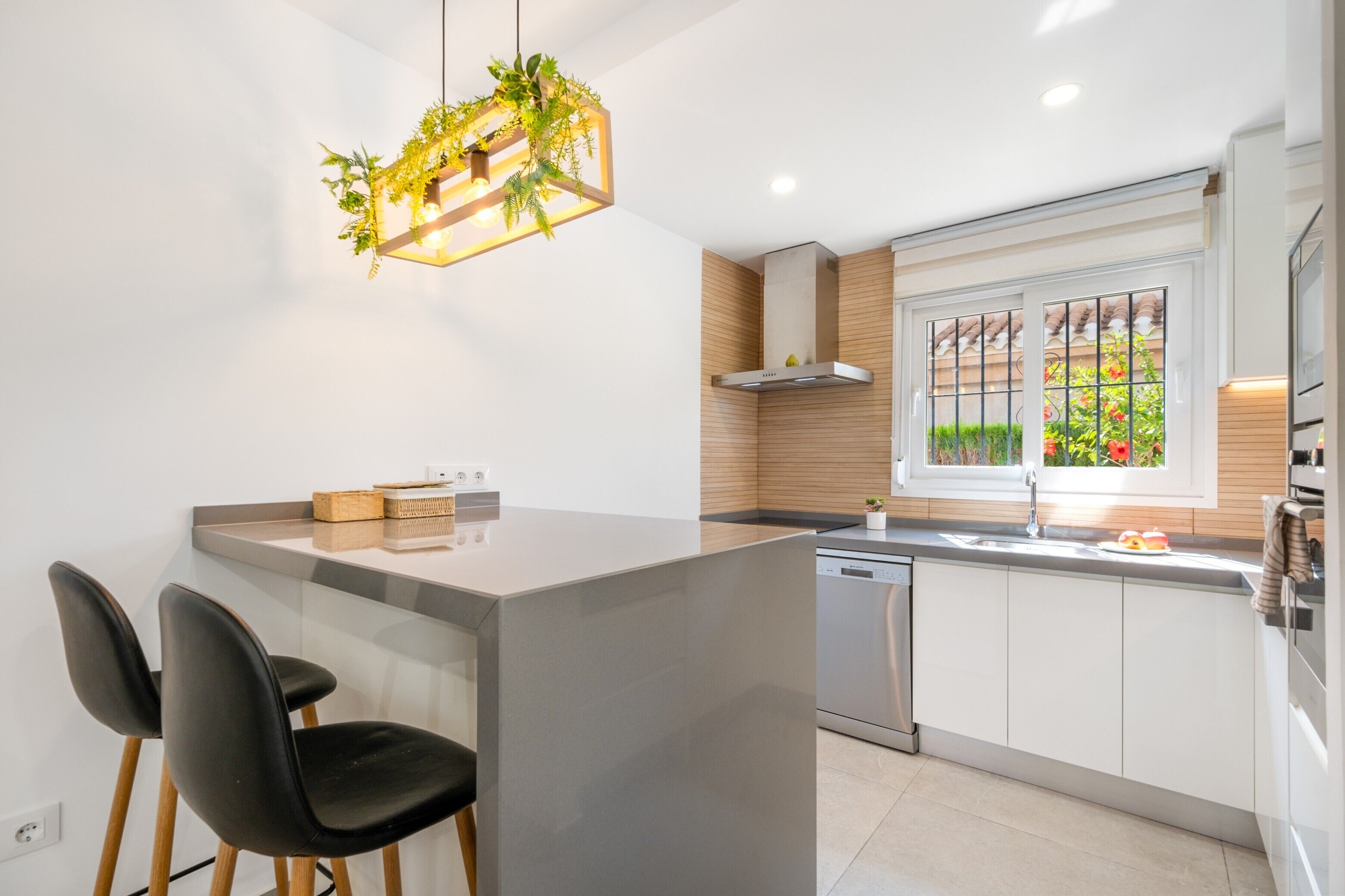 A modern kitchen with a central island, two black chairs, a hanging light fixture with plants, and a window with a view of greenery.