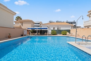 Une piscine dont le bord est revêtu de carreaux, un espace détente couvert et un palmier en arrière-plan.