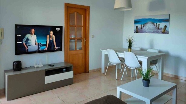 Una sala de estar con televisor, una puerta de madera, una zona de comedor con sillas, una mesa y un cuadro en la pared.