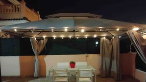 Un patio al aire libre con una mesa y sillas, una planta en maceta y un área cubierta con luces colgantes.