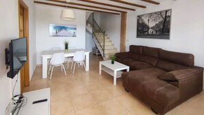 Una sala de estar con un sofá marrón, una mesa de comedor con sillas, un televisor y una escalera que sube a otro nivel.