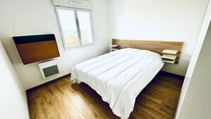 A bedroom with a bed, a window, a wall-mounted air conditioner, and a wooden floor.