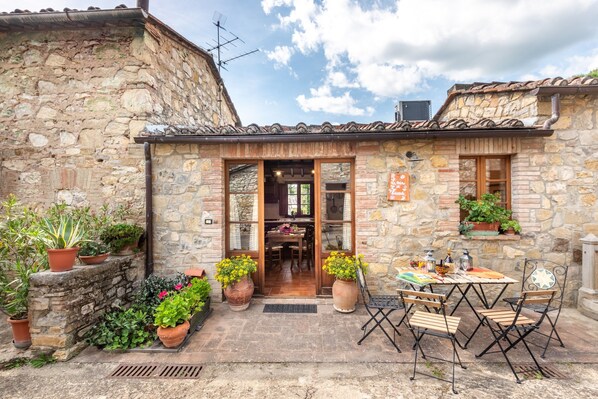 Un edificio de piedra con techo de tejas, puerta de madera y un pequeño patio con mesa y sillas.