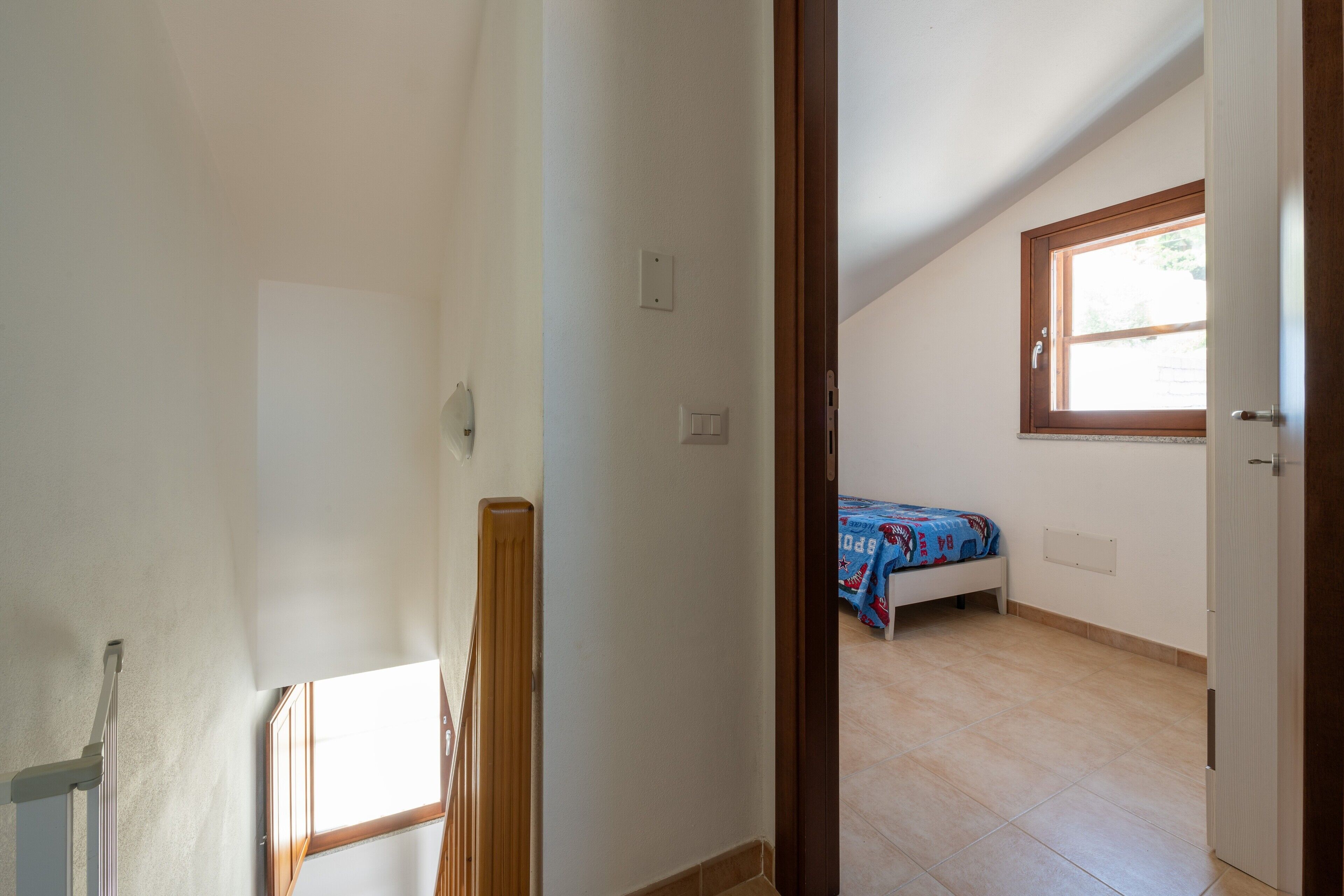 Ein Zimmer mit schrägem Dach, einem Fenster, einem Bett mit blauer Bettdecke und einer Tür.