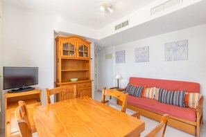 A room with a wooden dining table, chairs, a red sofa, a television on a stand, and a wooden cabinet with glass doors. - Apartment 'Primera Linea Calpe' with Sea View, Private Pool and Air Conditioning (Calpe)