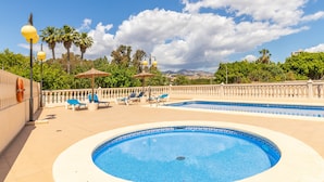 Zona de piscina con sillas de descanso y sombrillas, rodeada de una valla y palmeras.