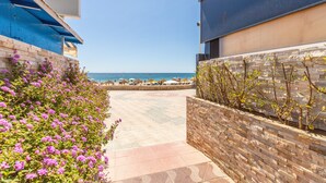 Un sendero que conduce a una playa con vista al mar, un edificio azul con toldo blanco y un muro de piedra con plantas.