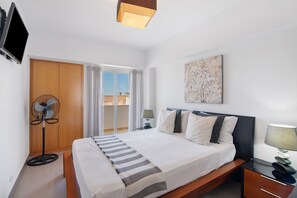 A hotel room with a bed, a standing fan, a TV, a window with curtains, and a view of buildings outside.