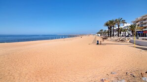Ein Strand mit blauem Himmel, sandigem Ufer und Gebäuden im Hintergrund.