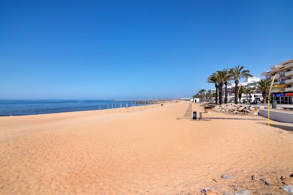 A beach with a clear sky, a sandy shore, and buildings in the background.