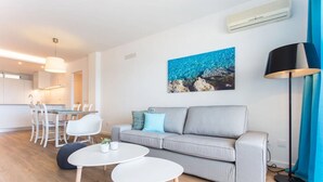 Una sala de estar moderna con un sofá gris, sillas blancas, una mesa de comedor y un cuadro de una costa rocosa.