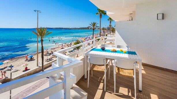Un balcón con vista a la playa y al océano, una mesa con mantel azul y una barandilla blanca.