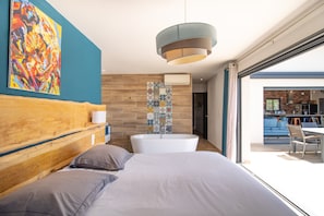 Een moderne slaapkamer met een houten hoofdeinde, een groot bed, een badkuip en een kleurrijk abstract schilderij aan de muur.