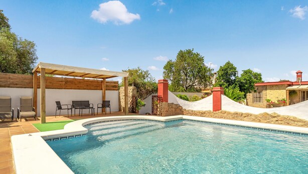 Ein Pool mit Pergola und Liegestühlen, umgeben von einer Steinmauer und Grünpflanzen.
