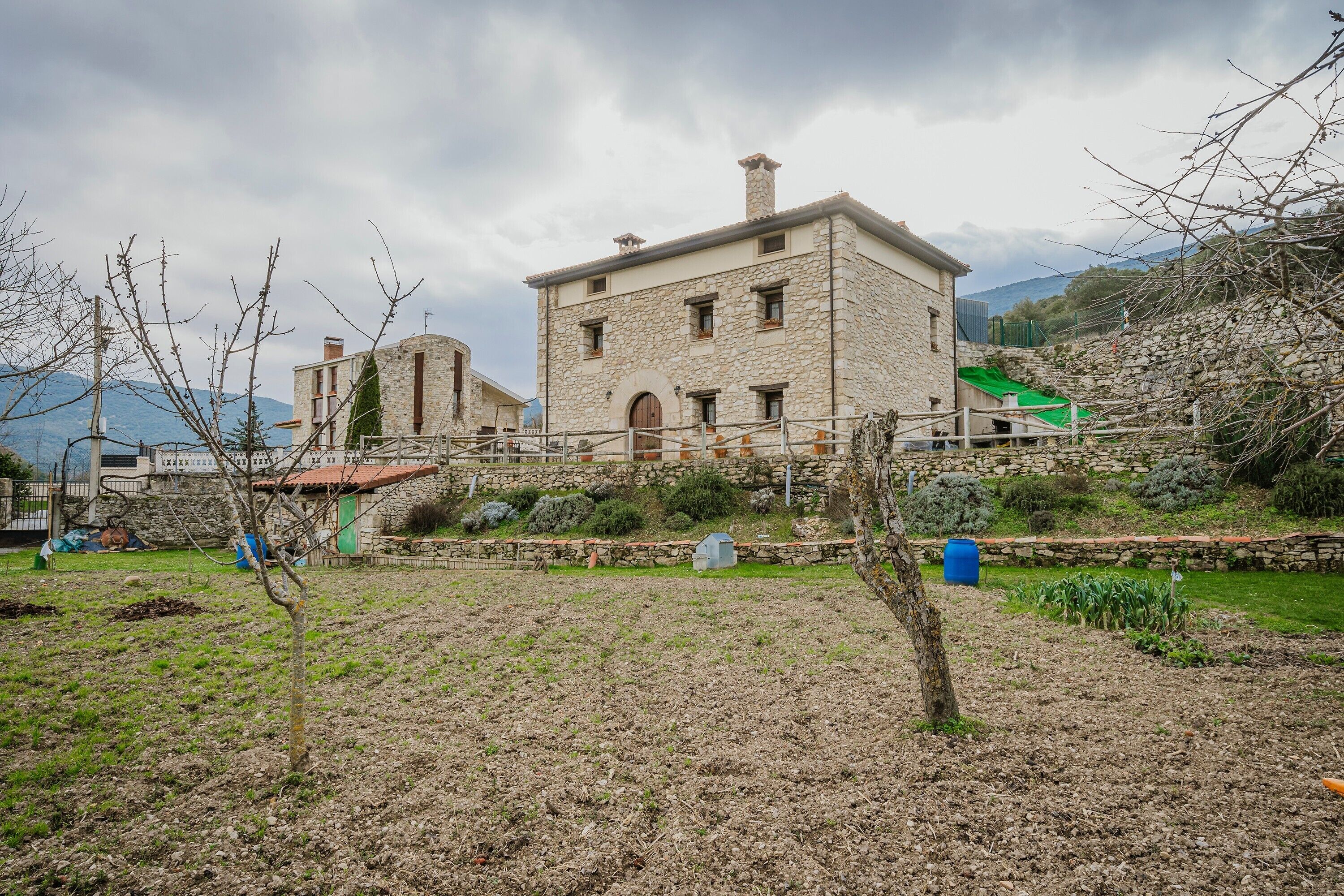 Un edificio de piedra con techo plano, un pequeño jardín y vista a montañas al fondo.