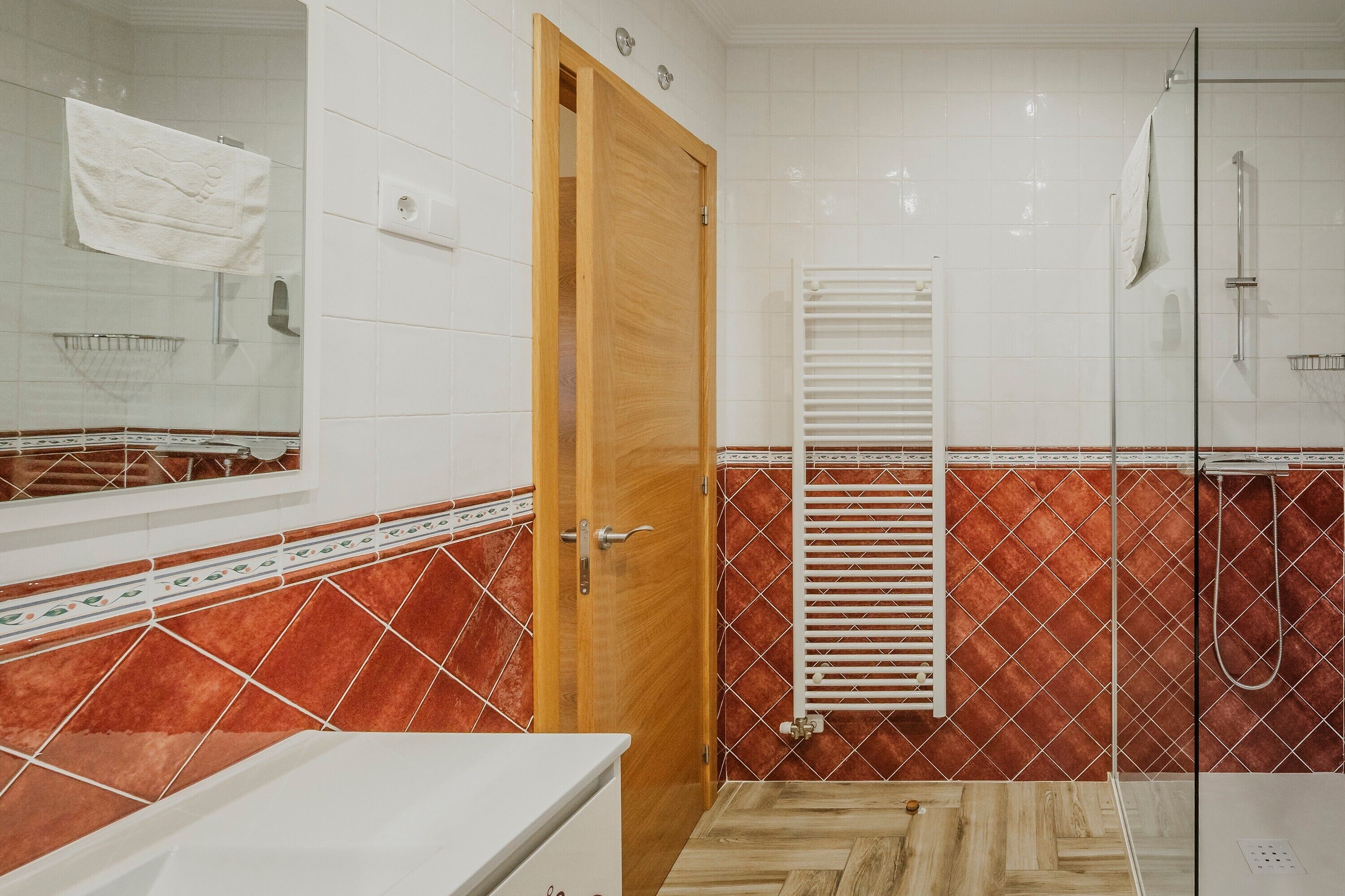 Un baño con puerta de madera, un lavamanos blanco, una ducha con puerta de vidrio y un revestimiento de pared con diseño rojo.