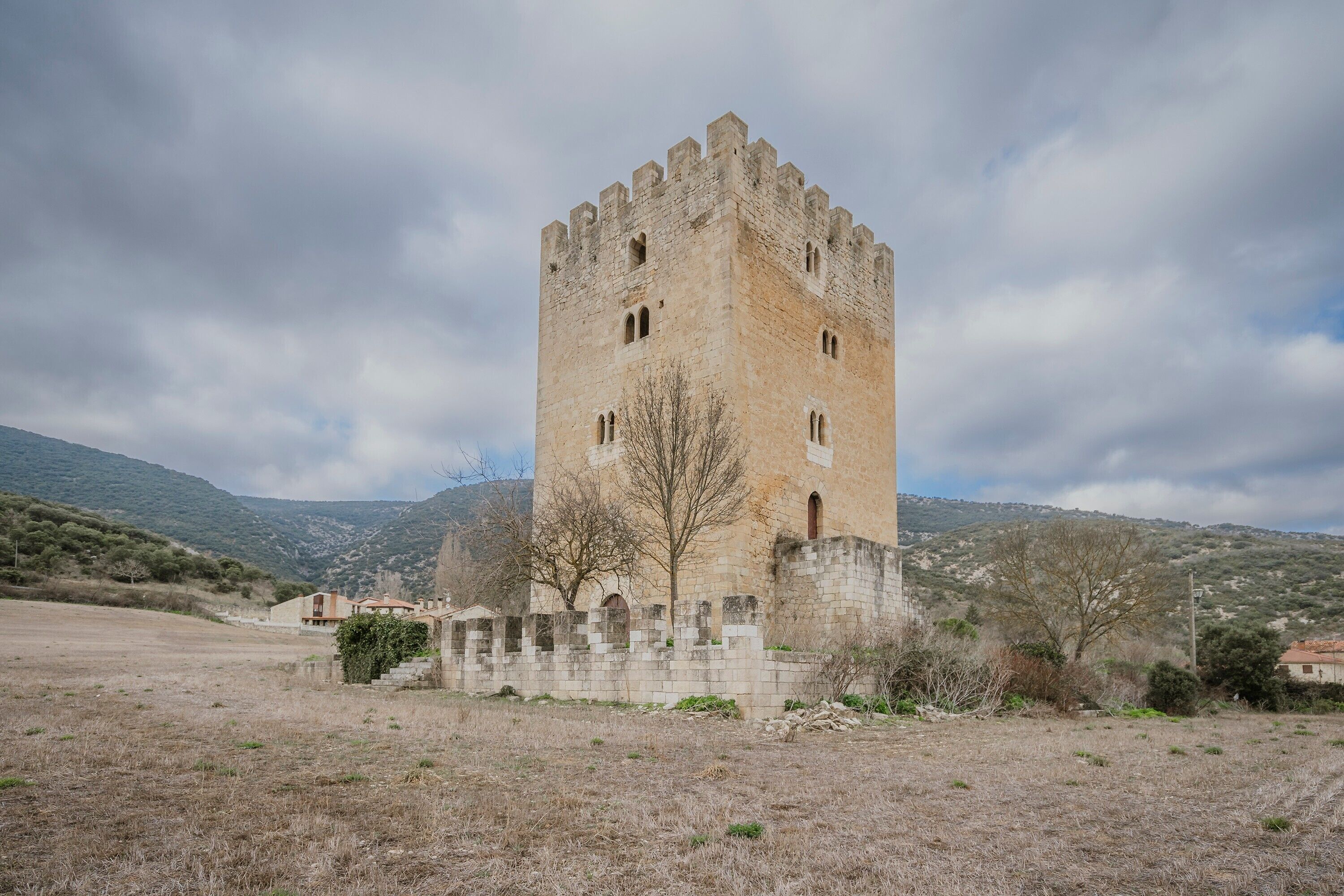 Una torre de piedra histórica con ventanas de arco, rodeada por un campo seco y colinas distantes.