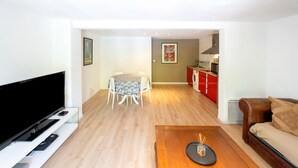 A living room with a flat-screen TV, a wooden coffee table, a sofa, a dining table with chairs, and a kitchen area with red cabinets. - Apartment 'Appartement Dans Maison Urcuit' with Mountain View, Private Pool and Wi-Fi (Urcuit)