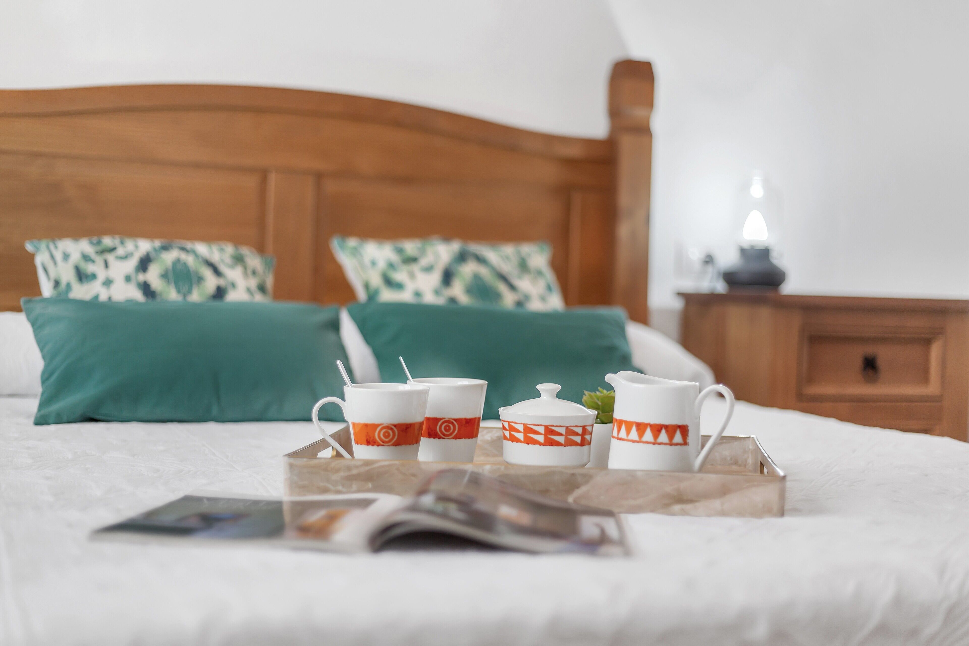 A neatly made bed with a tray of cups and a teapot, a magazine, and a wooden nightstand with a lamp.