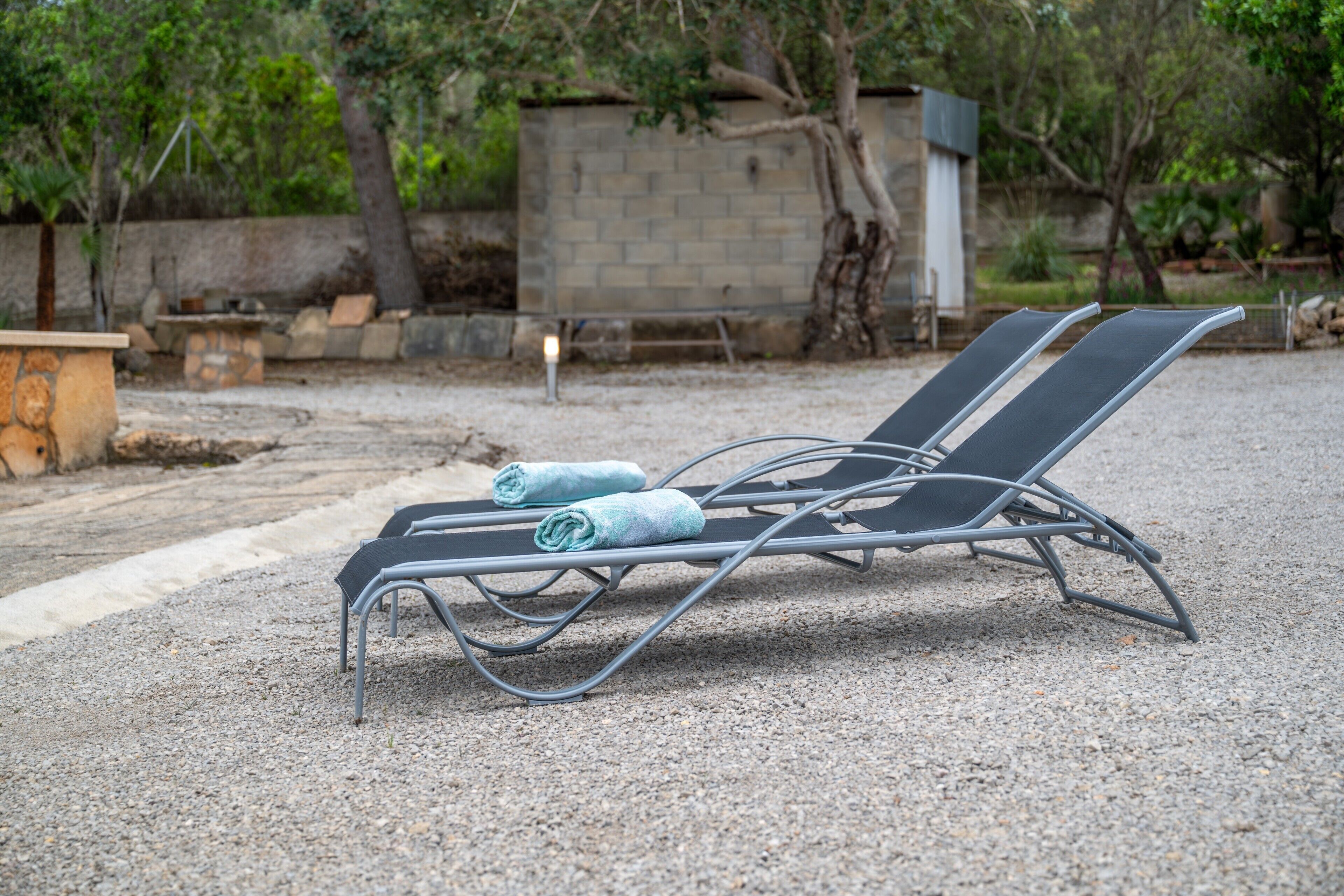 Une chaise longue en métal, avec des serviettes, posée sur du gravier, avec un mur de pierre et des arbres en arrière-plan.