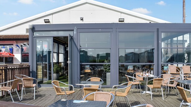 An outdoor patio area with wicker chairs and tables, a wooden deck, and a building with large windows and a white roof. - 3 bedroom accommodation in Heysham, near Lancaster (Heysham, near Lancaster)
