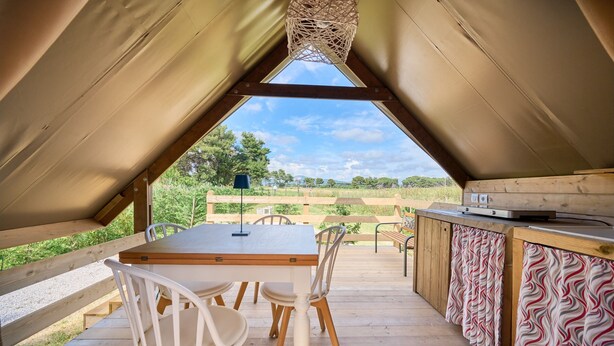 Une tente dotée d’une terrasse en bois, d’une table avec des chaises, d’un luminaire suspendu et offrant une vue sur l’extérieur.