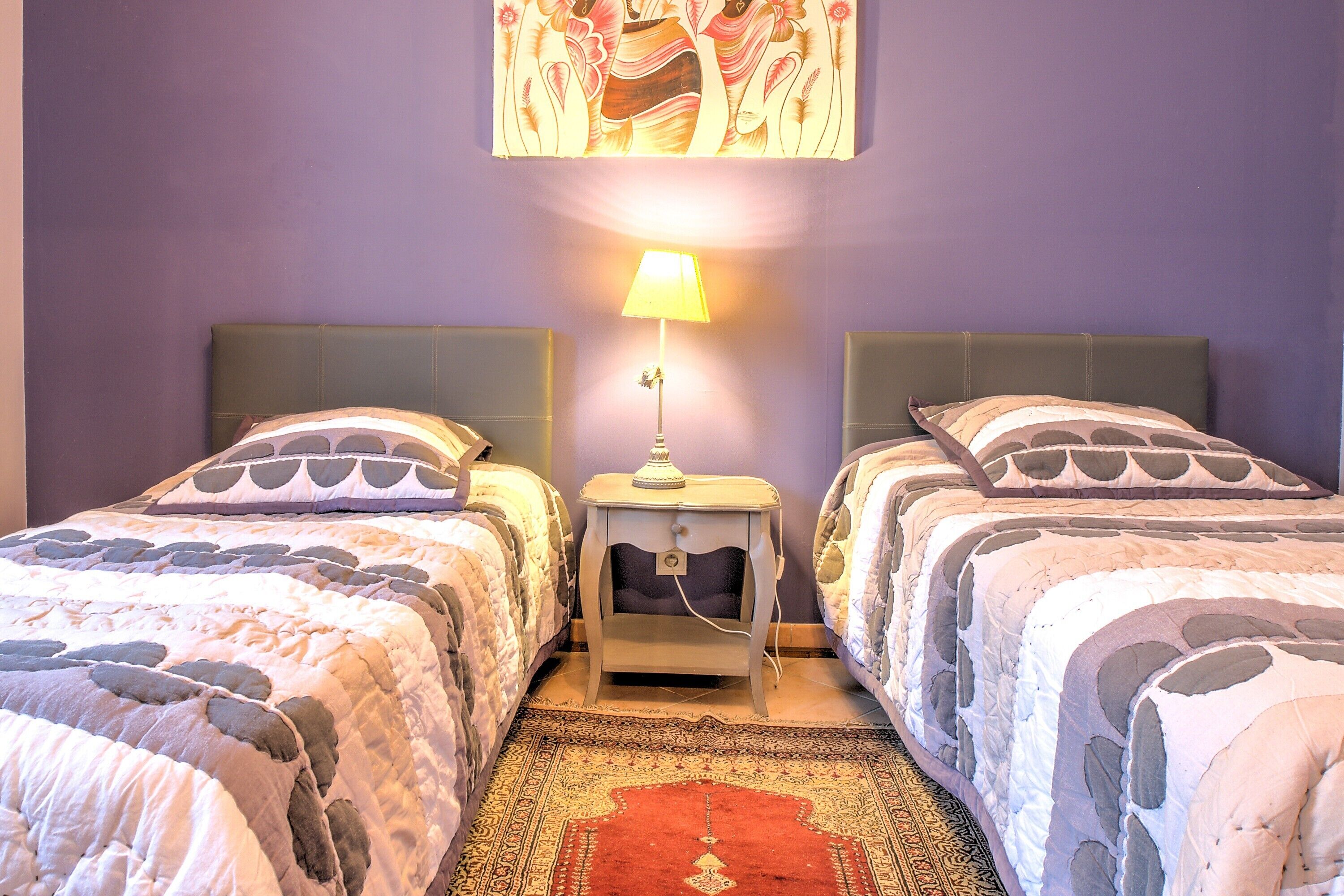 Une chambre avec deux lits, une table de chevet avec une lampe, un tapis à motifs et un tableau au mur.
