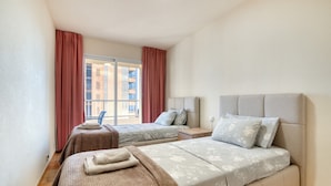 A hotel room with two beds, a window with curtains, a view of buildings, and a chair.