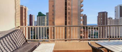 A balcony with a sofa, table, and chairs, overlooking a cityscape with tall buildings.
